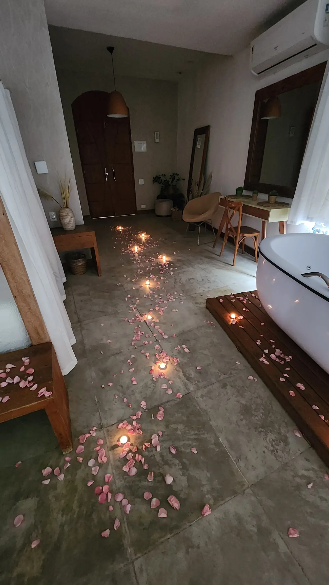 Corredor interno com banheira, cadeira, mesa com espelho e caminho de velas e pétalas de rosa no chão.