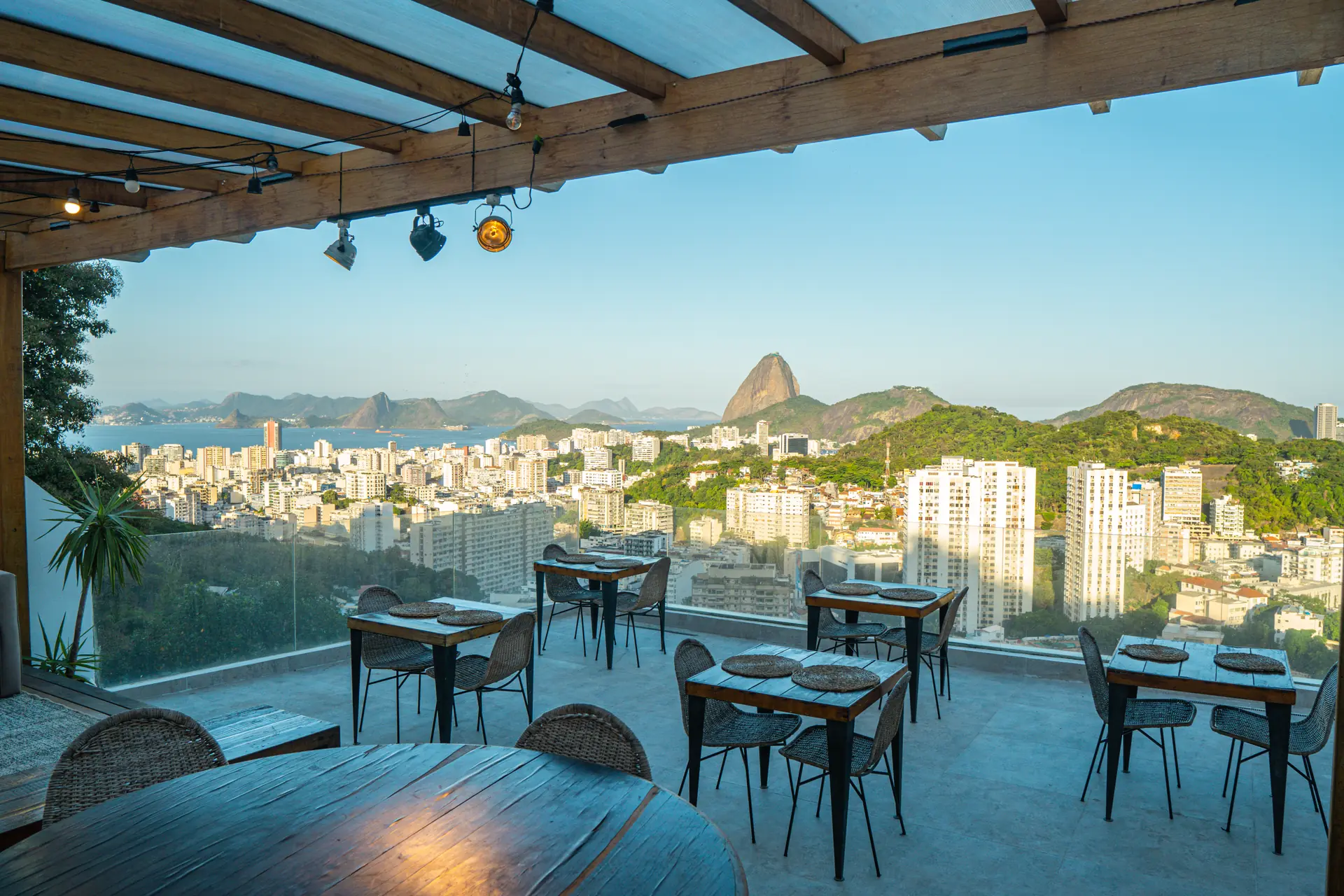 Rooftop do Santa Vista Rio ao entardecer: mesas sob a pérgola de madeira e a cidade ao fundo
