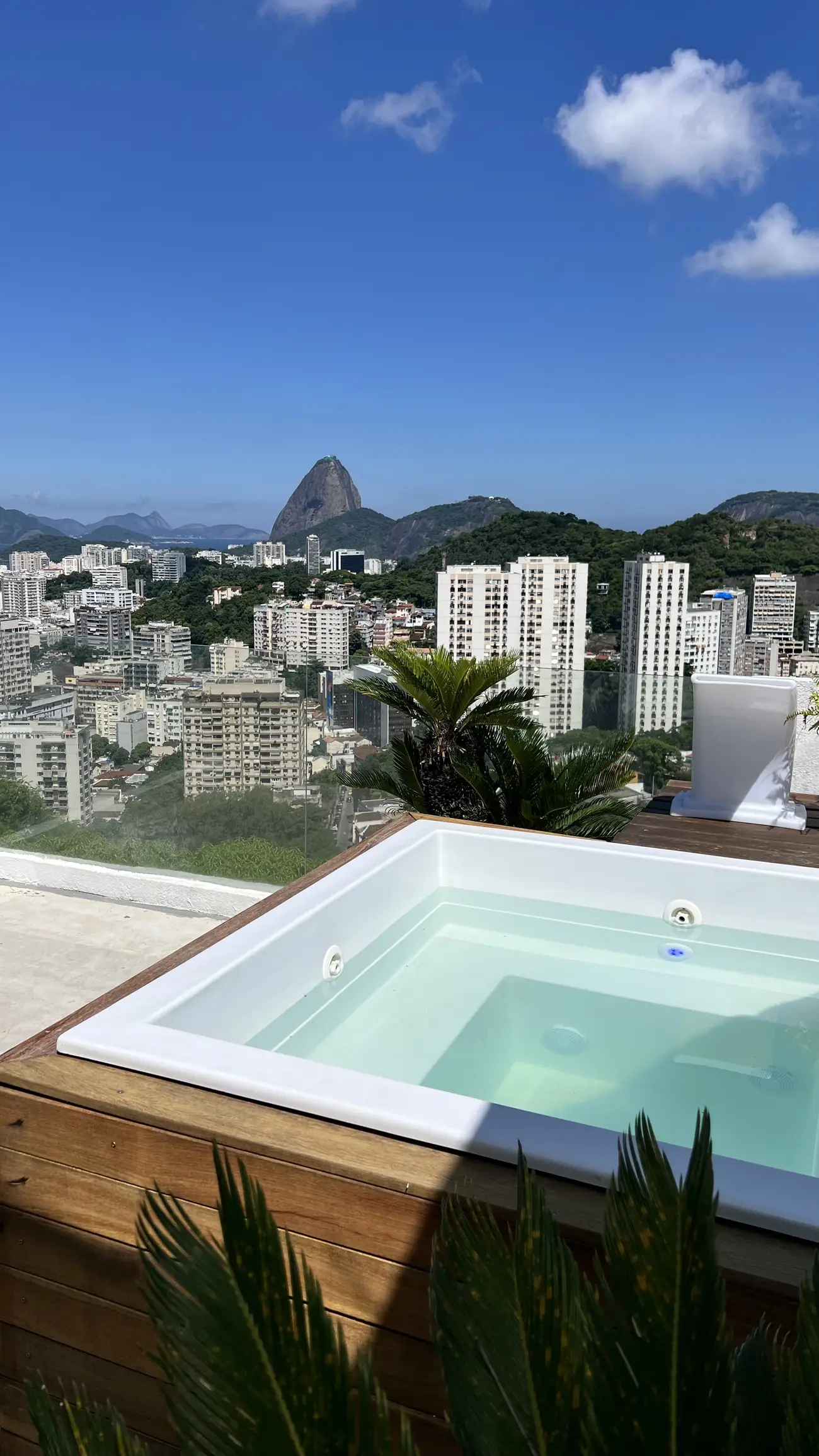 Ofurô com água limpa em deck de madeira, com vista para prédios e o Pão de Açúcar ao fundo sob céu azul.