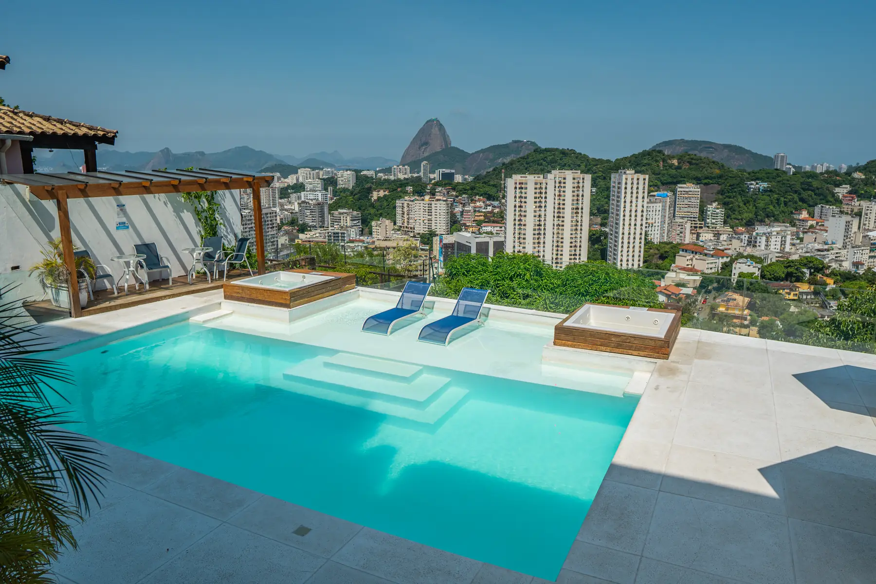 Piscina com duas espreguiçadeiras dentro da água, dois ofurôs ao lado e uma área com mesa e cadeiras sob pergolado, com vista para o Pão de Açúcar e prédios ao fundo.