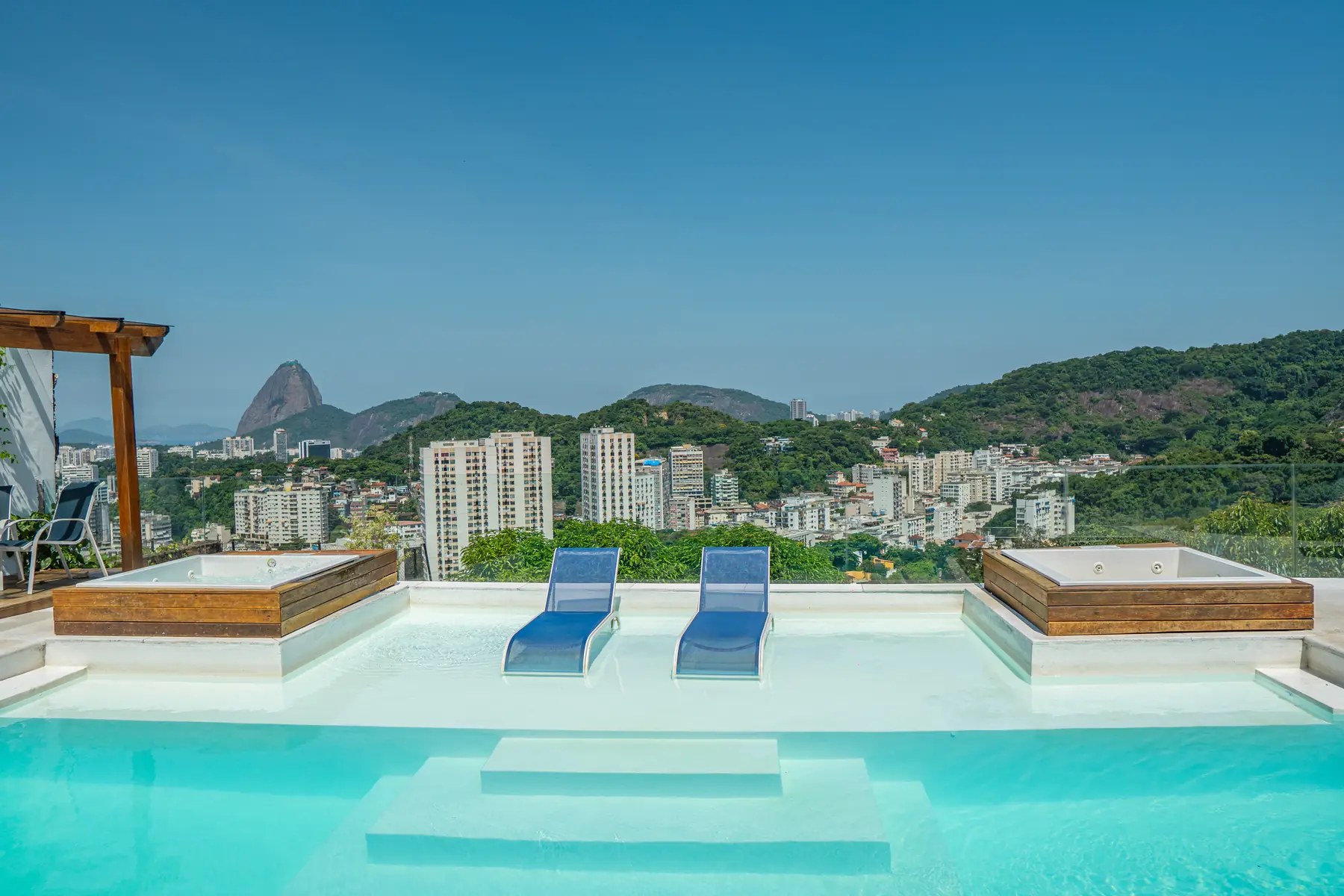 Piscina com duas espreguiçadeiras dentro da água e dois ofurôs de madeira nas laterais, com vista para prédios e morros ao fundo.