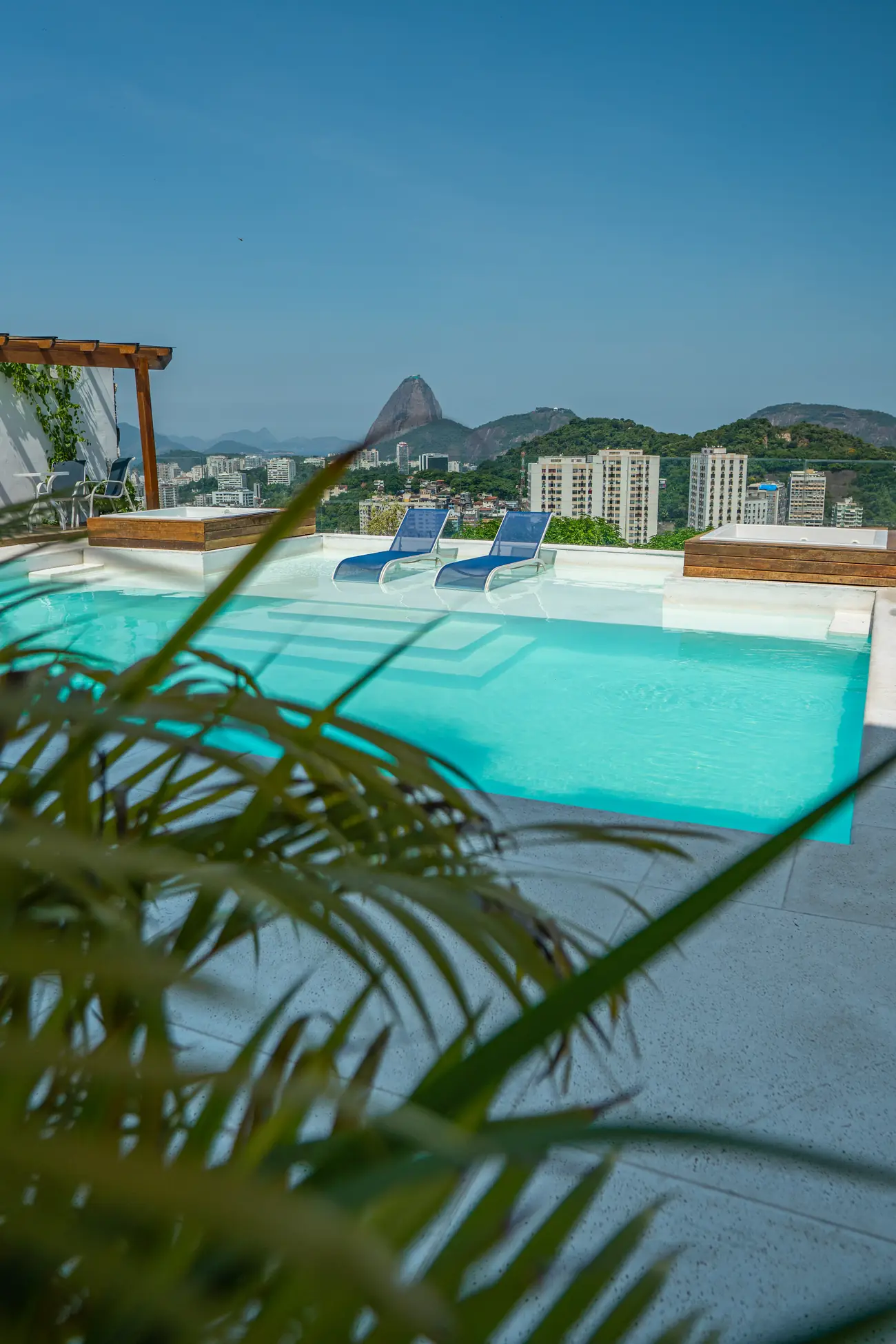 Piscina com duas espreguiçadeiras azuis dentro da água, com vista para o Pão de Açúcar e prédios ao fundo, sob céu azul.