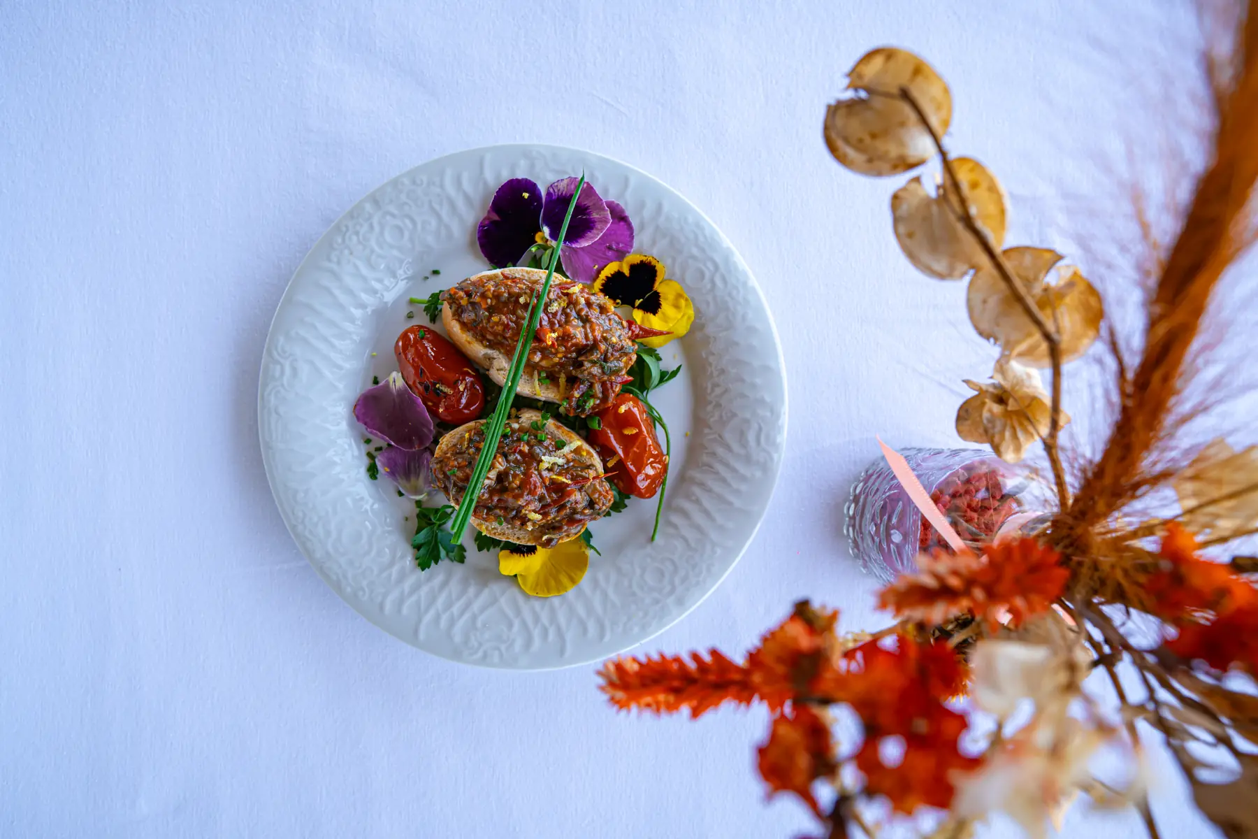 Prato com duas porções de bruschetta com tomate e ervas, decorado com flores comestíveis, sobre toalha branca, com arranjo floral desfocado ao lado.