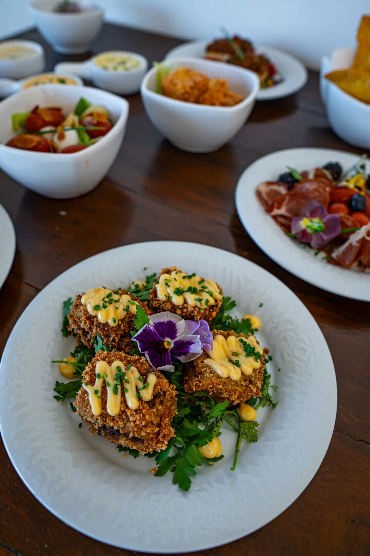 Prato em primeiro plano com quatro croquetes empanados decorados com molho e uma flor comestível roxa, dispostos sobre folhas verdes. Ao fundo, outros pratos e bowls com petiscos e saladas estão distribuídos sobre uma mesa de madeira.
