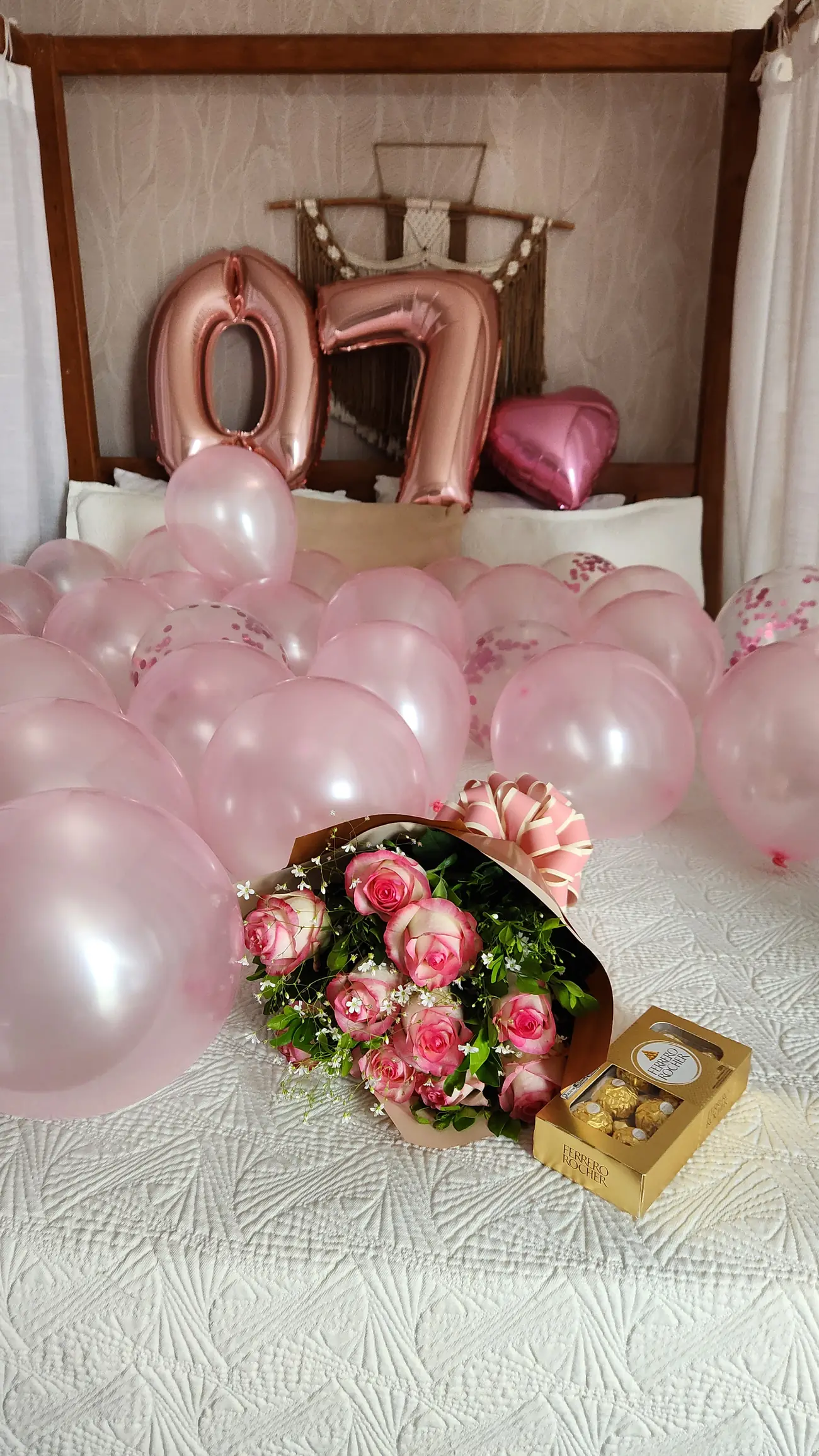 Cama com balões cor de rosa, um buquê de rosas e uma caixa de chocolates sobre colcha branca.