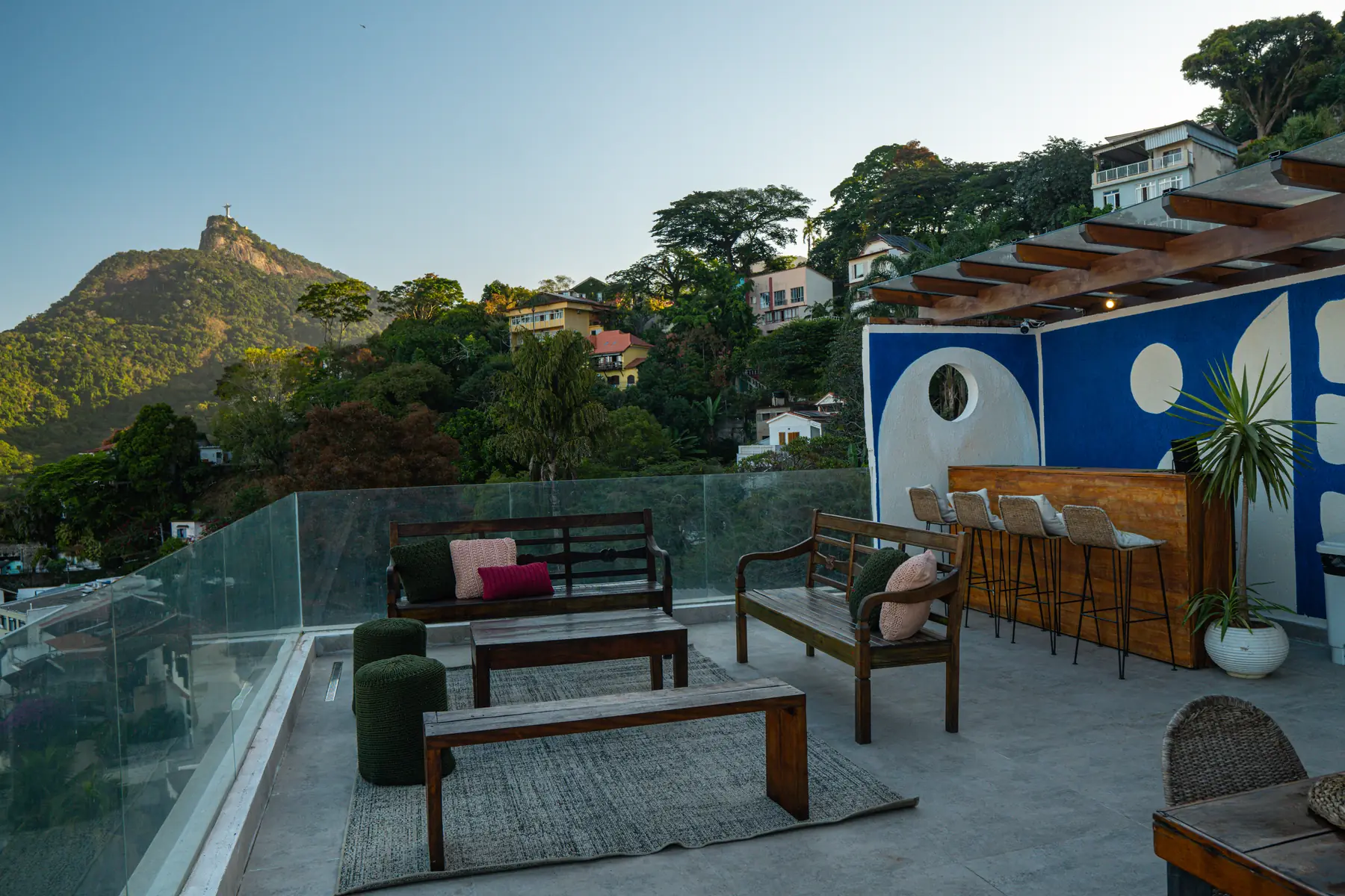 Área externa com bancos de madeira, almofadas, pufes e um bar com banquetas, com vista para o Cristo Redentor e vegetação ao redor.
