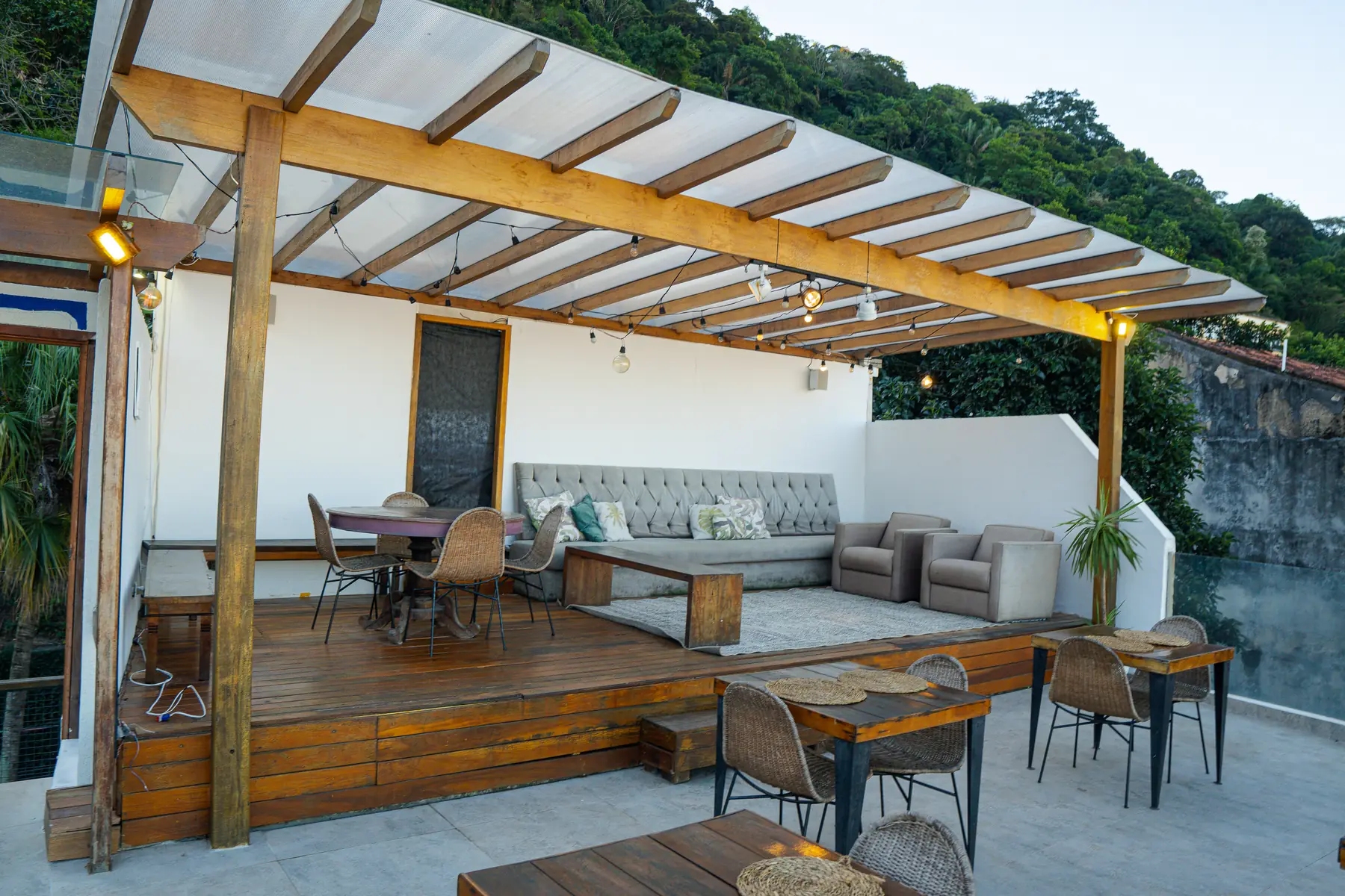 Área externa com deck de madeira coberto por estrutura com vigas e iluminação, contendo sofá, poltronas, mesas e cadeiras.