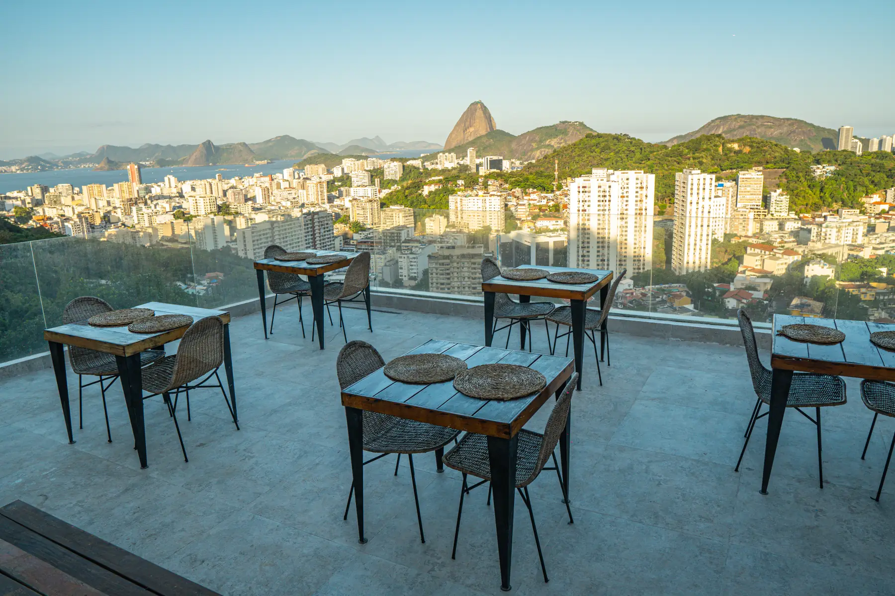 Área externa com mesas e cadeiras dispostas em um rooftop com vista para a cidade do Rio de Janeiro e o Pão de Açúcar ao fundo.