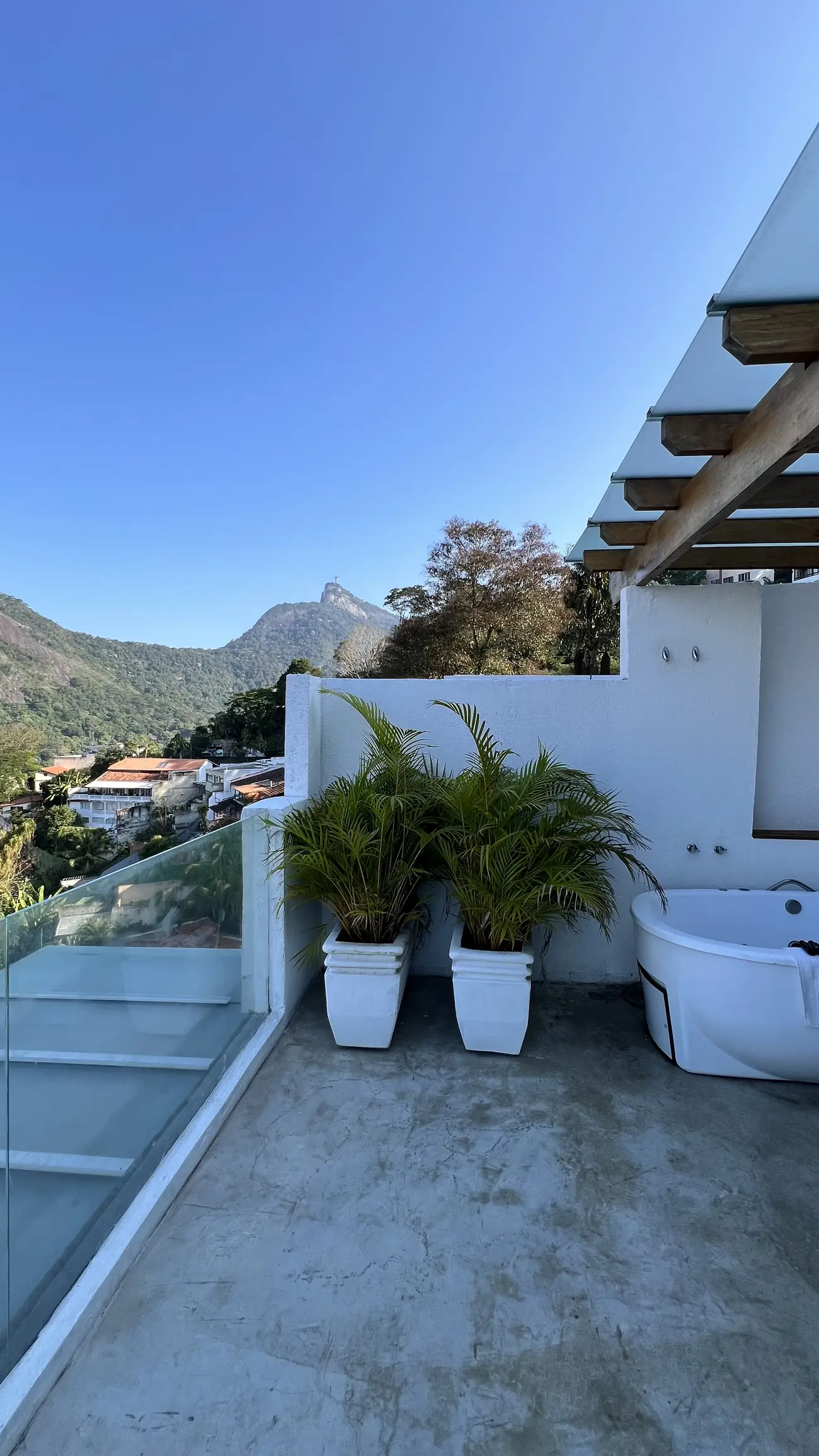 Varanda externa com duas plantas em vasos brancos, banheira de hidromassagem e vista para o Cristo Redentor e morros ao fundo.